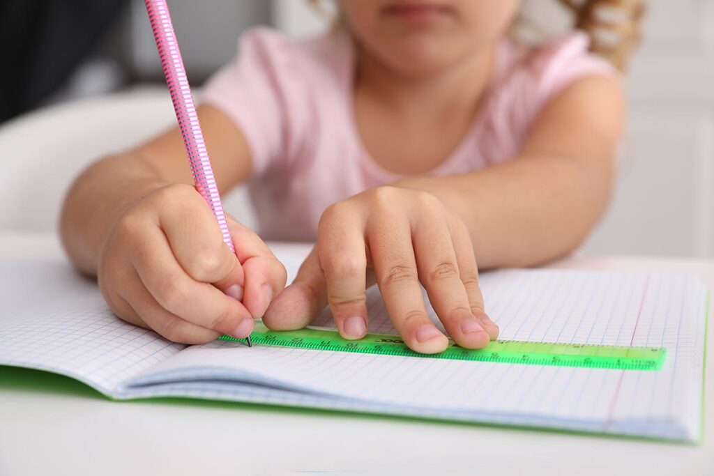 A young girl sits at a table, focused on writing in her notebook with a pencil, surrounded by colorful stationery.