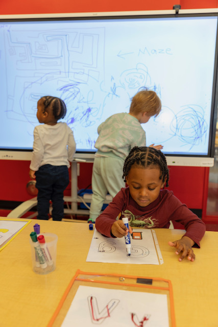 A group of children gathered around a table, drawing on a whiteboard with colorful markers.