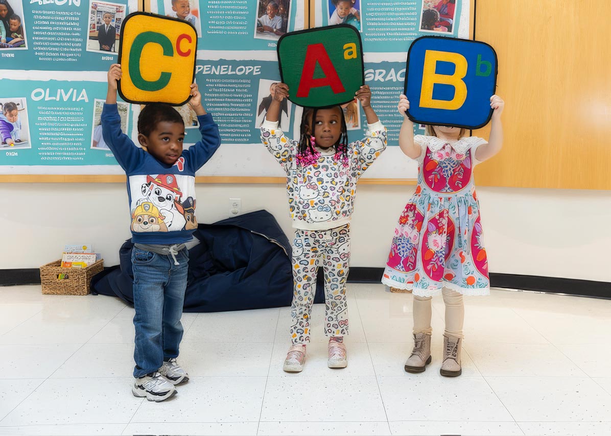 Three smiling children in a classroom hold up large letters, participating in an interactive learning activity.