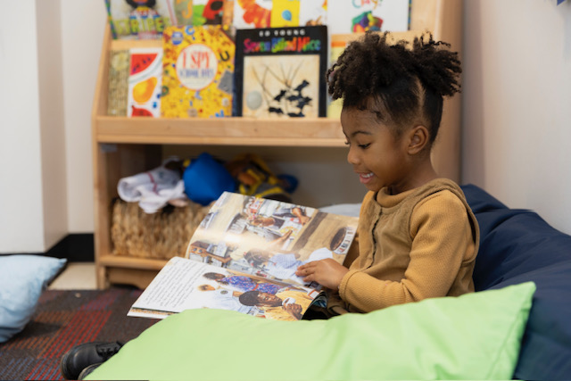 A young girl sits at a desk in a classroom, engrossed in reading a book.