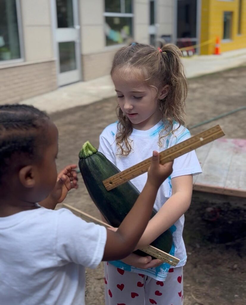 Two children proudly hold a large cucumber in front of a building, smiling and enjoying their harvest.